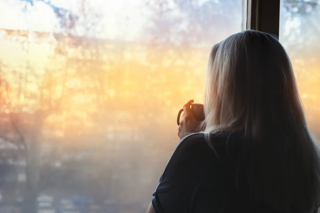 Blonde woman in deep thought standing by a window, with coffee cup in her hands, looking out the window. Hopeful.
