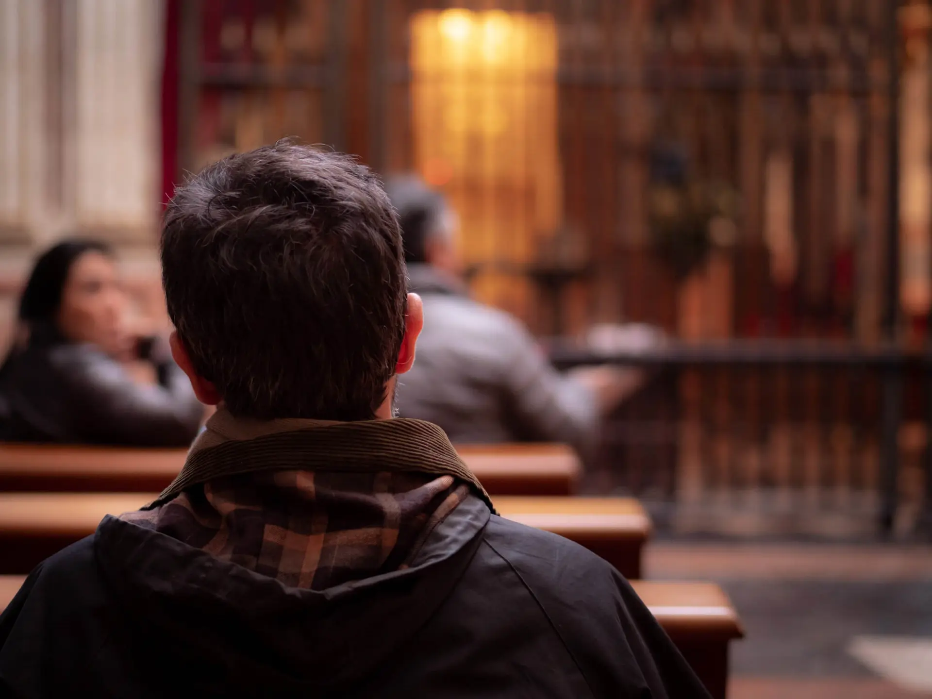 a person sits on a pew in a religious house of worship with a brown plaid and corduroy shirt facing the altar