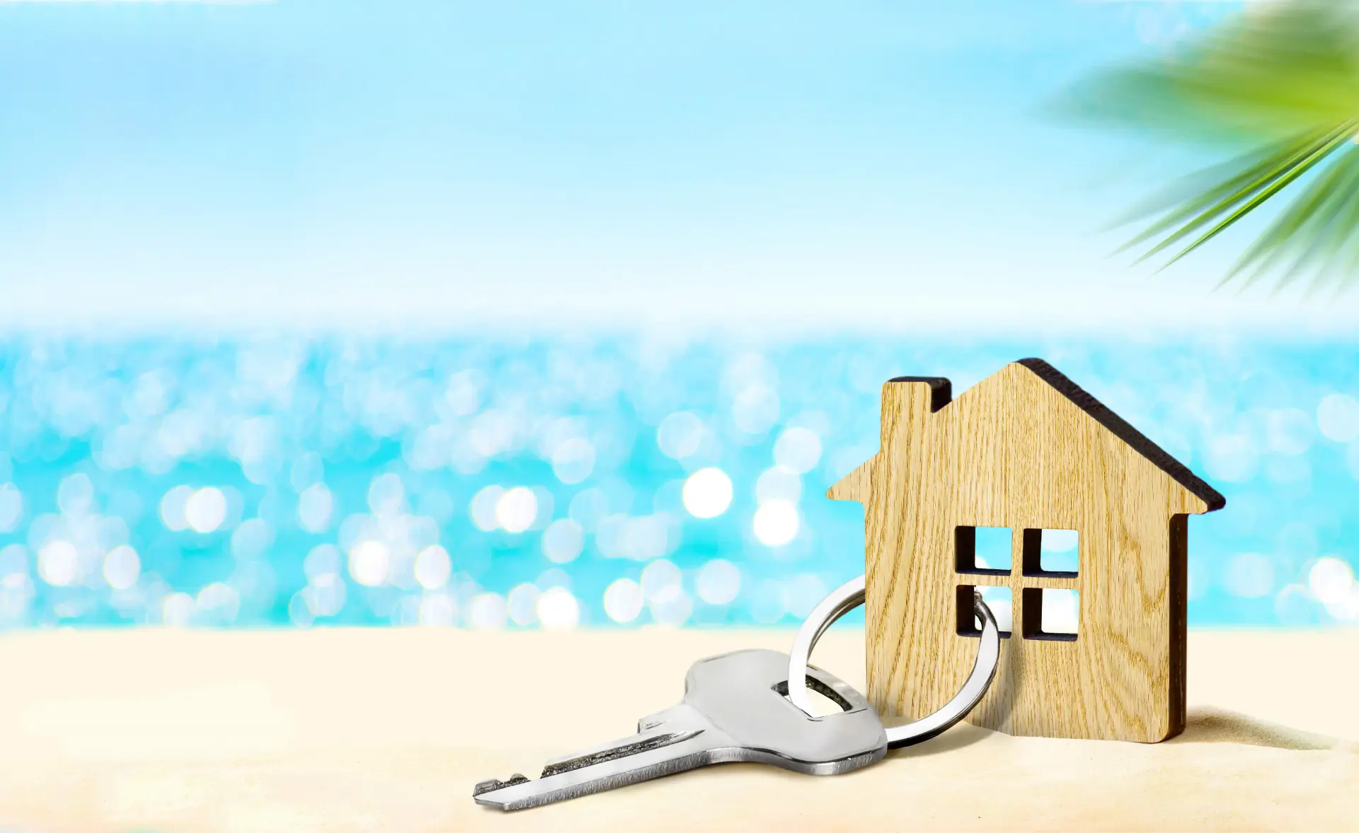 A flat wooden house keychain with a silver key attached by a key ring sits on a sandy beach with sparkling blue water in the background. There is a leafy palm in the top right hand corner.