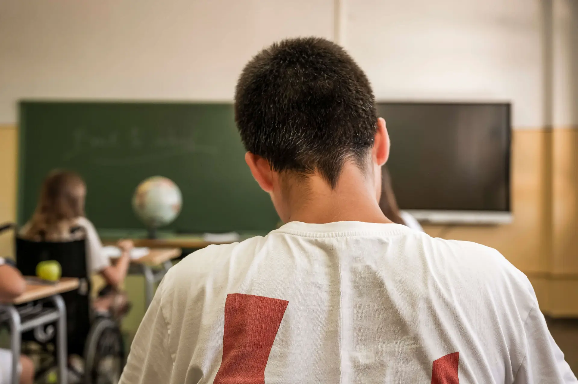 a distracted student in a white and red t shirt sits with their head facing down in a classroom