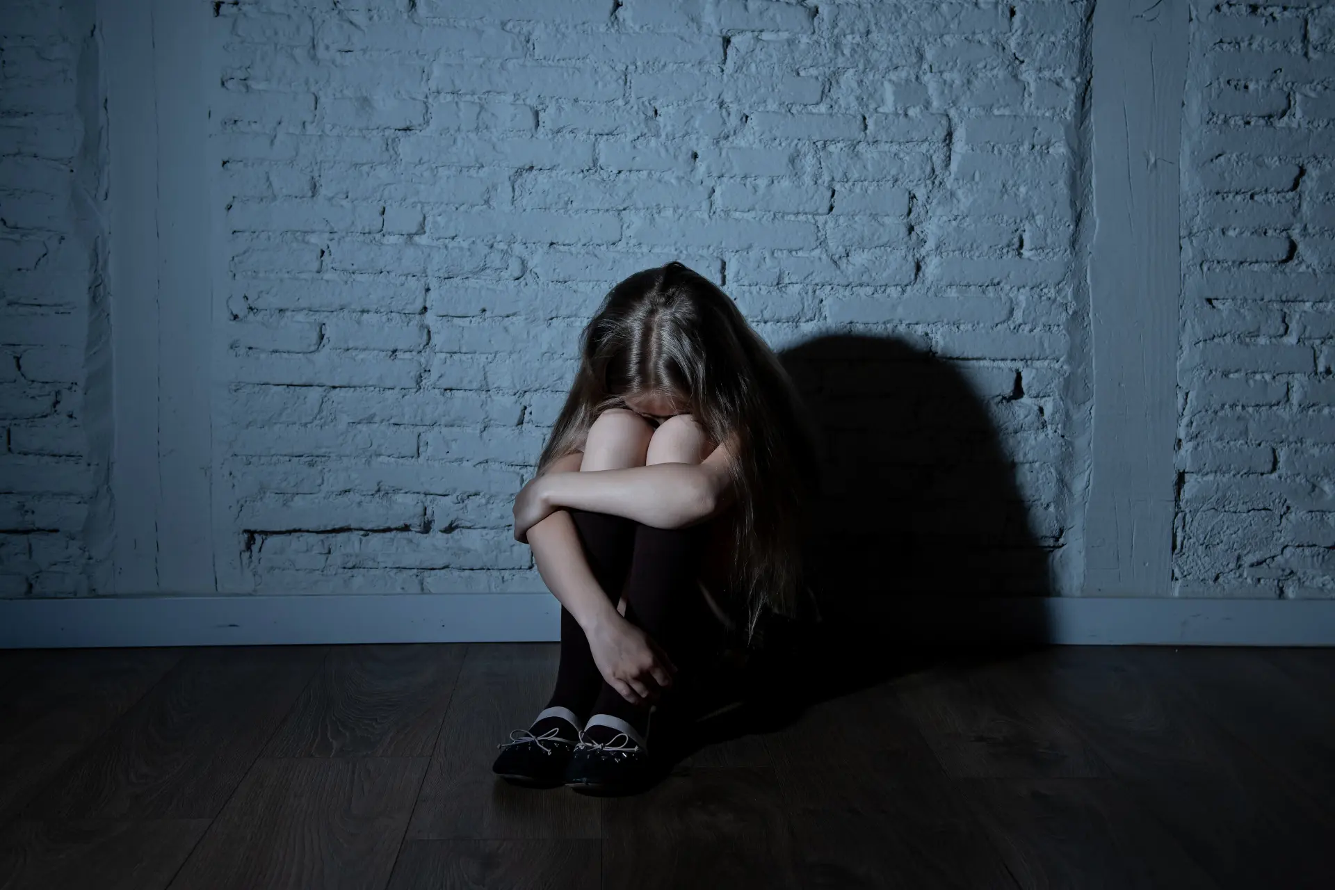 a young girl who may be suffering from abuse sits on the floor of a dark room with a white brick wall