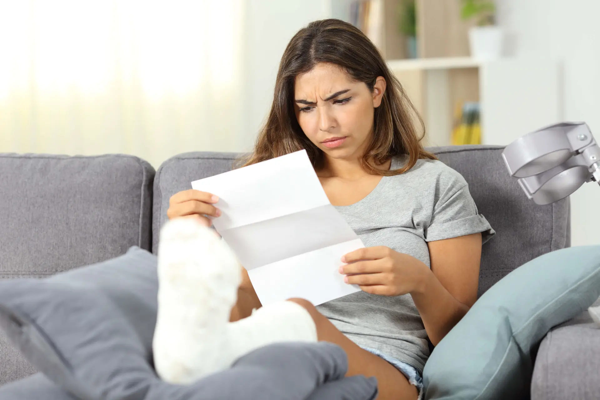 a woman in a grey t shirt reads a document while sitting on her couch with an injured and bandaged foot propped up on a pillow
