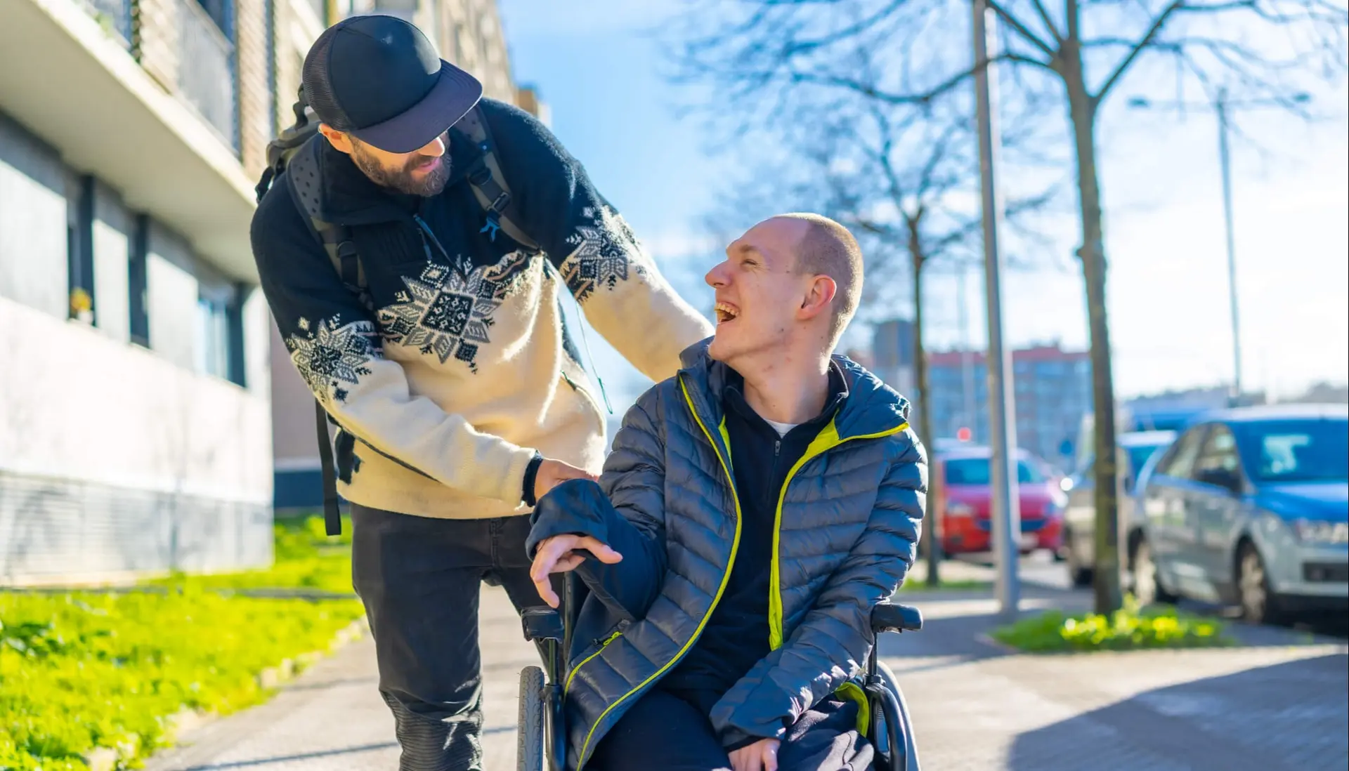a man in a wheelchair laughs at something his friend is saying to him while they're out on a sunny day