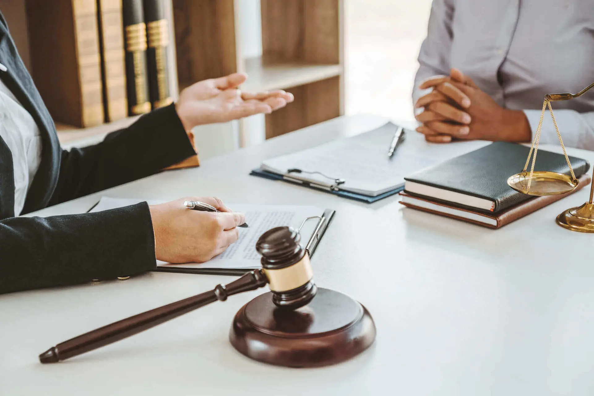 Two people sitting across from each other at a table. The person on the left holds their left hand out explaining something while the right one holds a pen over a document. The person on the right sits with their hands clasped together on the table.