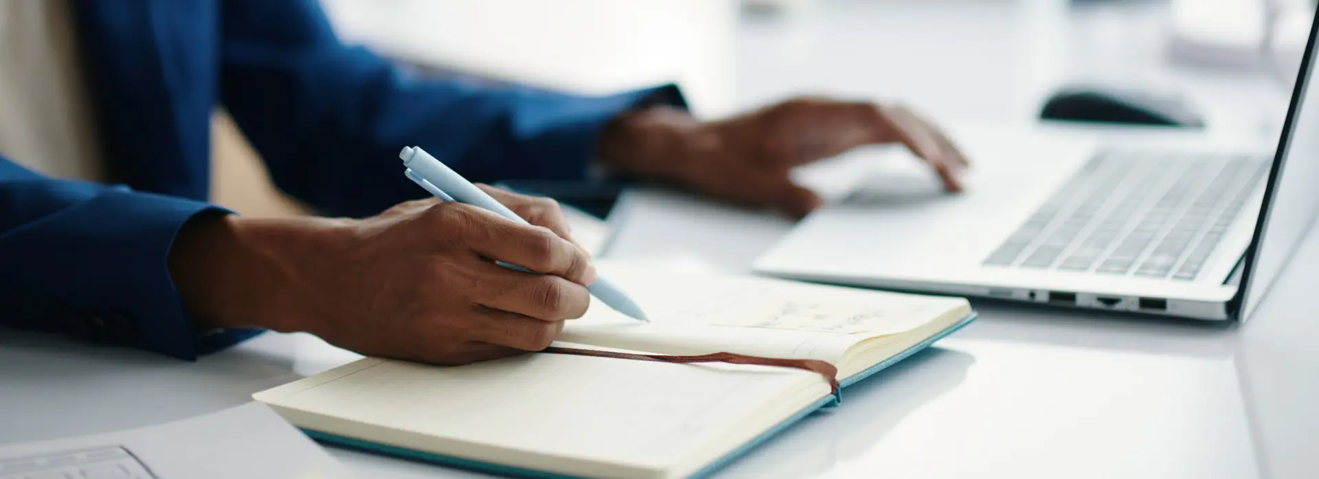 Person seated a table writing in a notebook with an open laptop near.