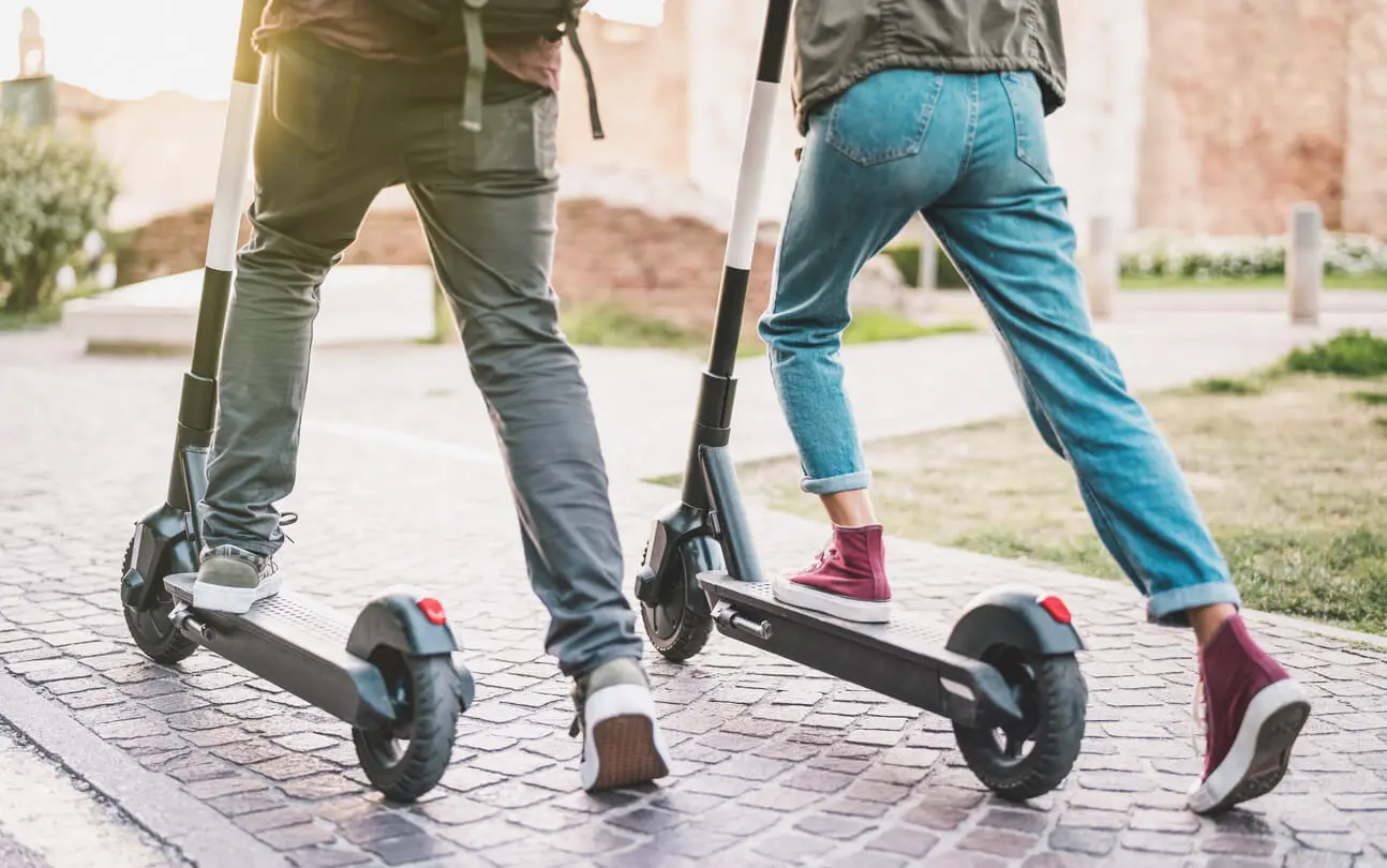 two people ride e-scooters on a brick path in a public park