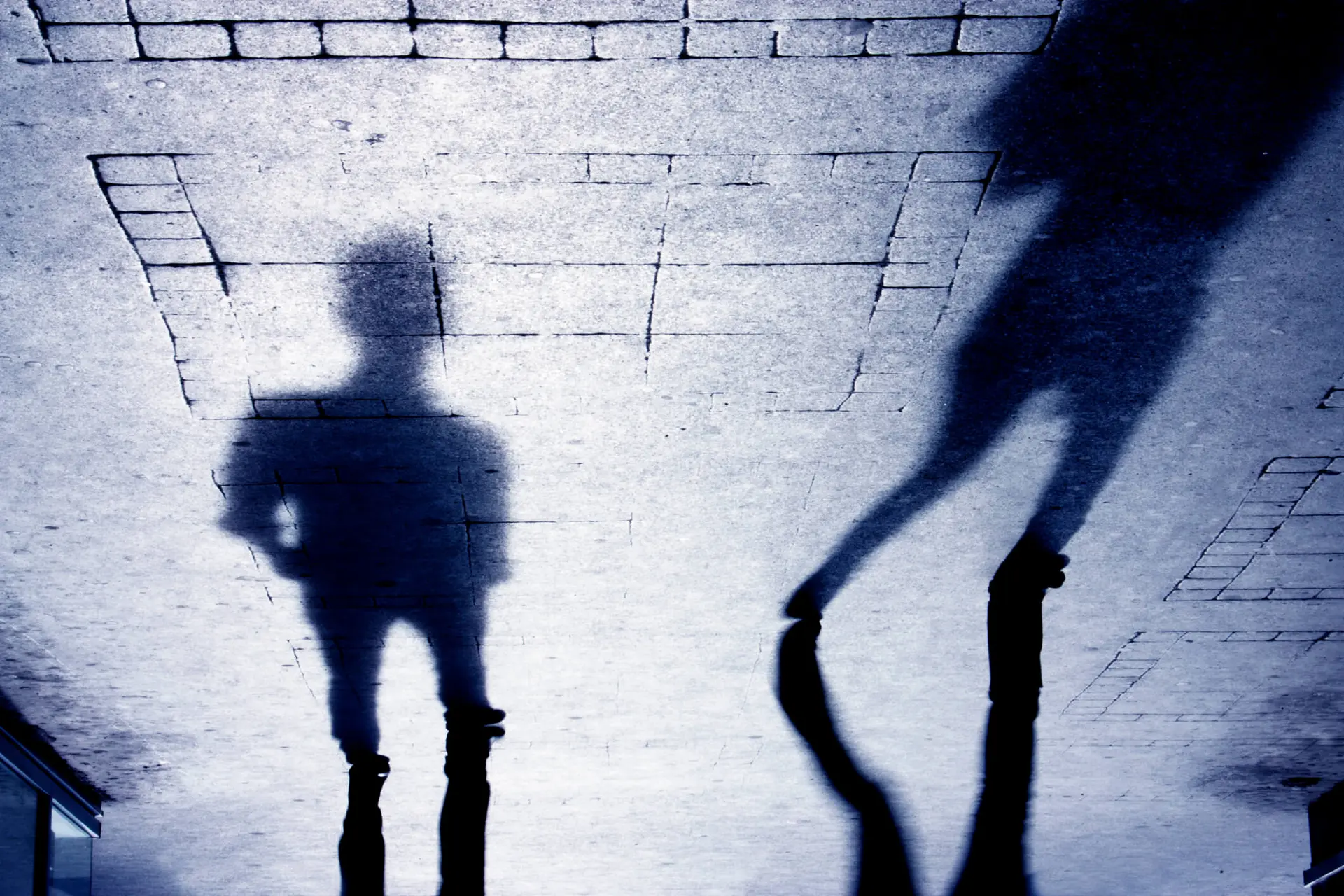 Two shadows of two people. The person on the left is standing with their hands on their hips, the person on the right is walking away from the person on the left.