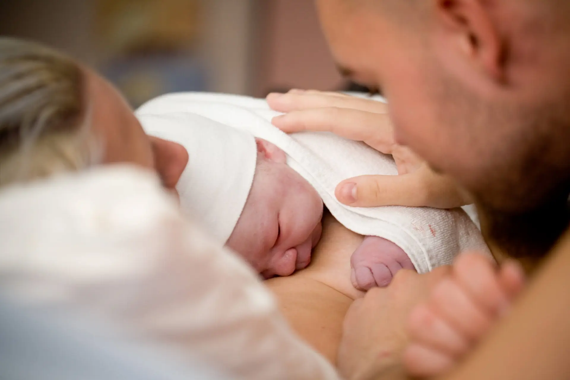 New born baby lays on their mother's chest with a white hat on and a white blanket wrapped around him. His father has his hand on the baby's back.