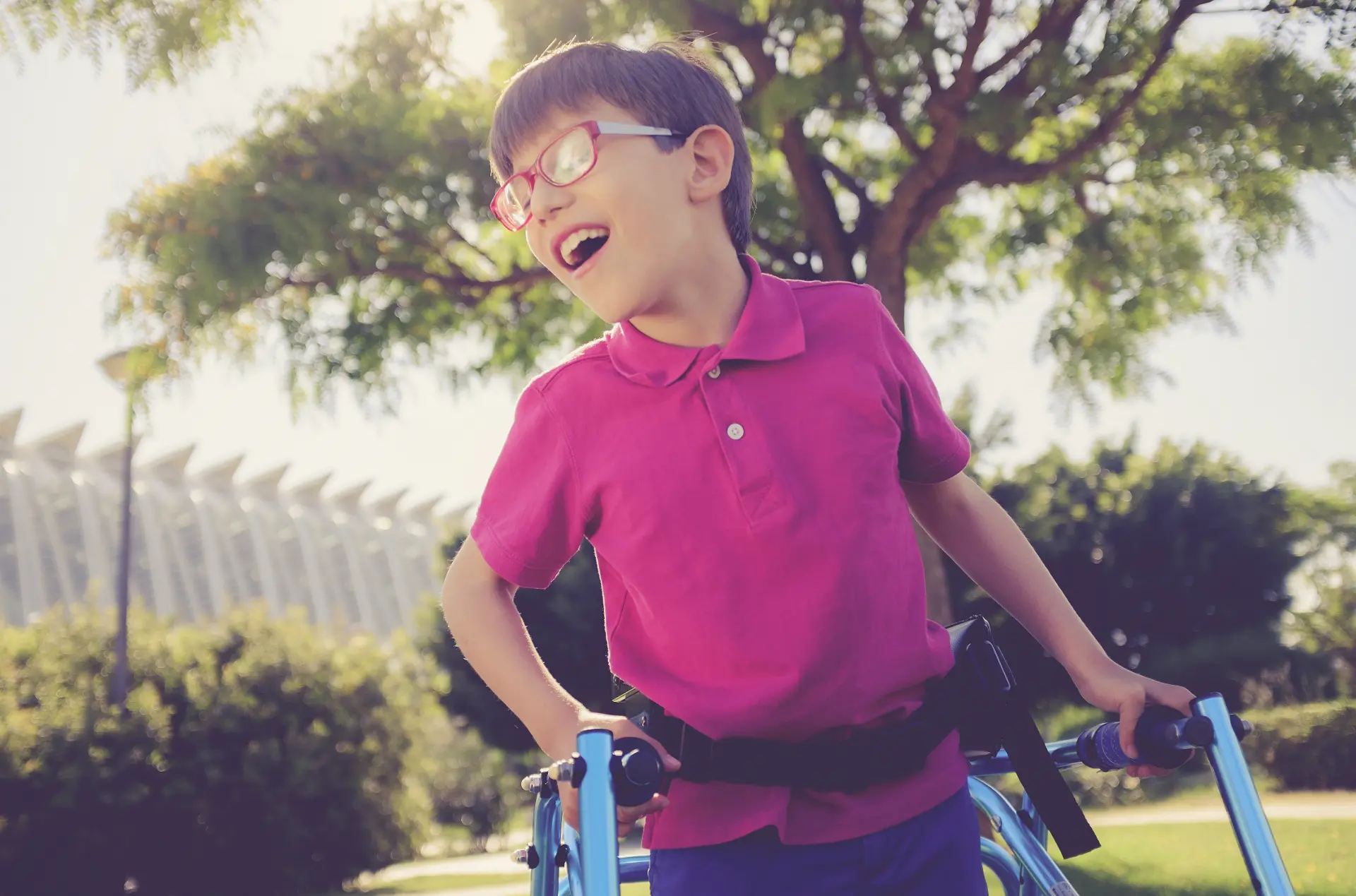 a child who may have cerebral palsy uses a blue walker to assist them in walking around a park