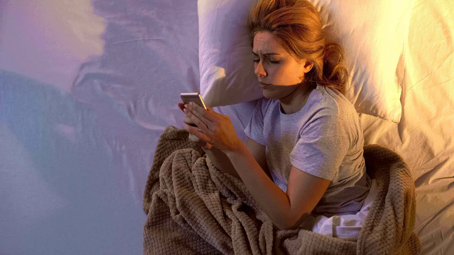 Woman laying in bed with a blanket on her. She is on her side while looking on her phone.