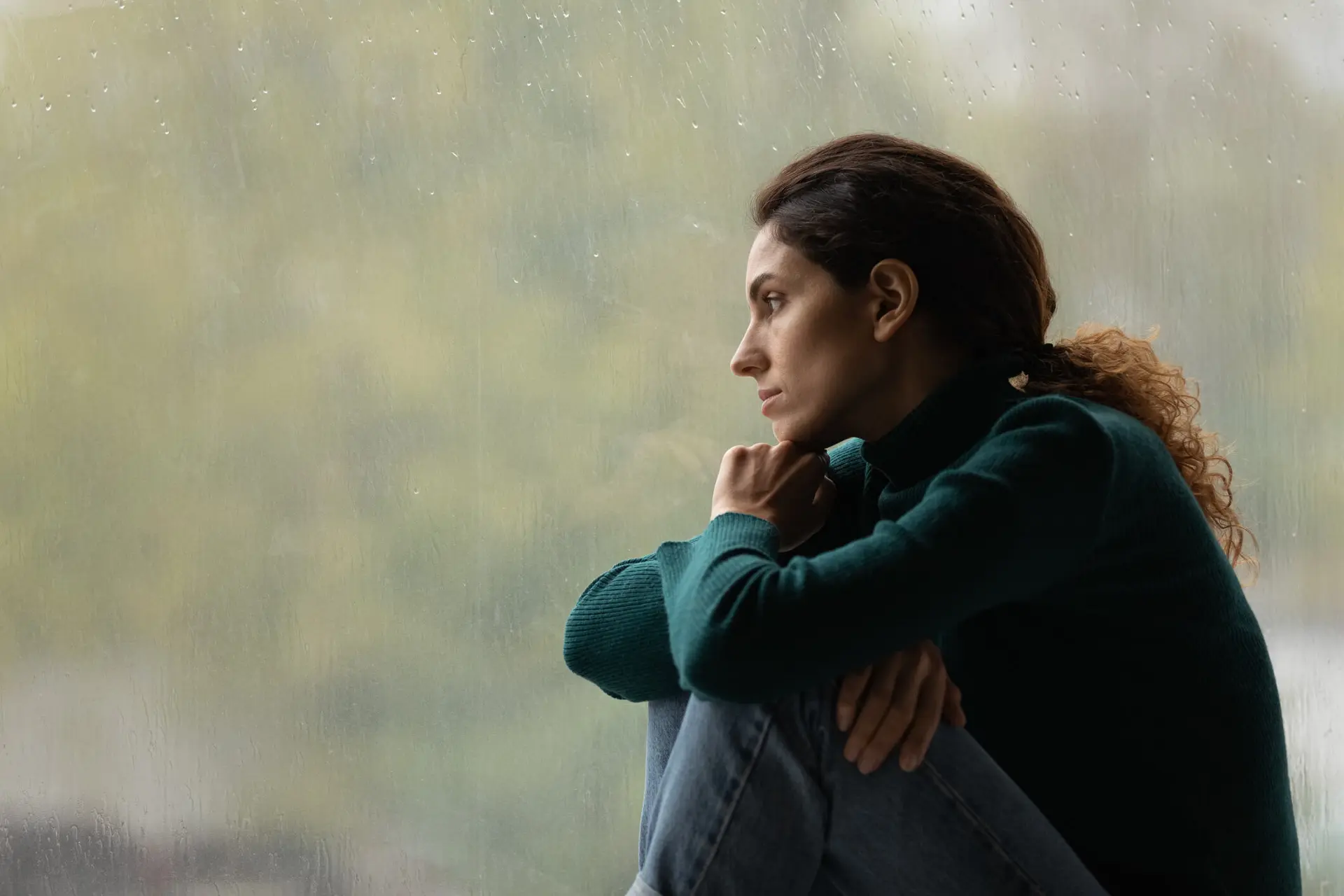 a woman in a green sweater who has possible been sexually abused or assaulted looks out a window contemplating her next steps a woman in a green sweater who has possible been sexually abused or assaulted looks out a window contemplating her next steps