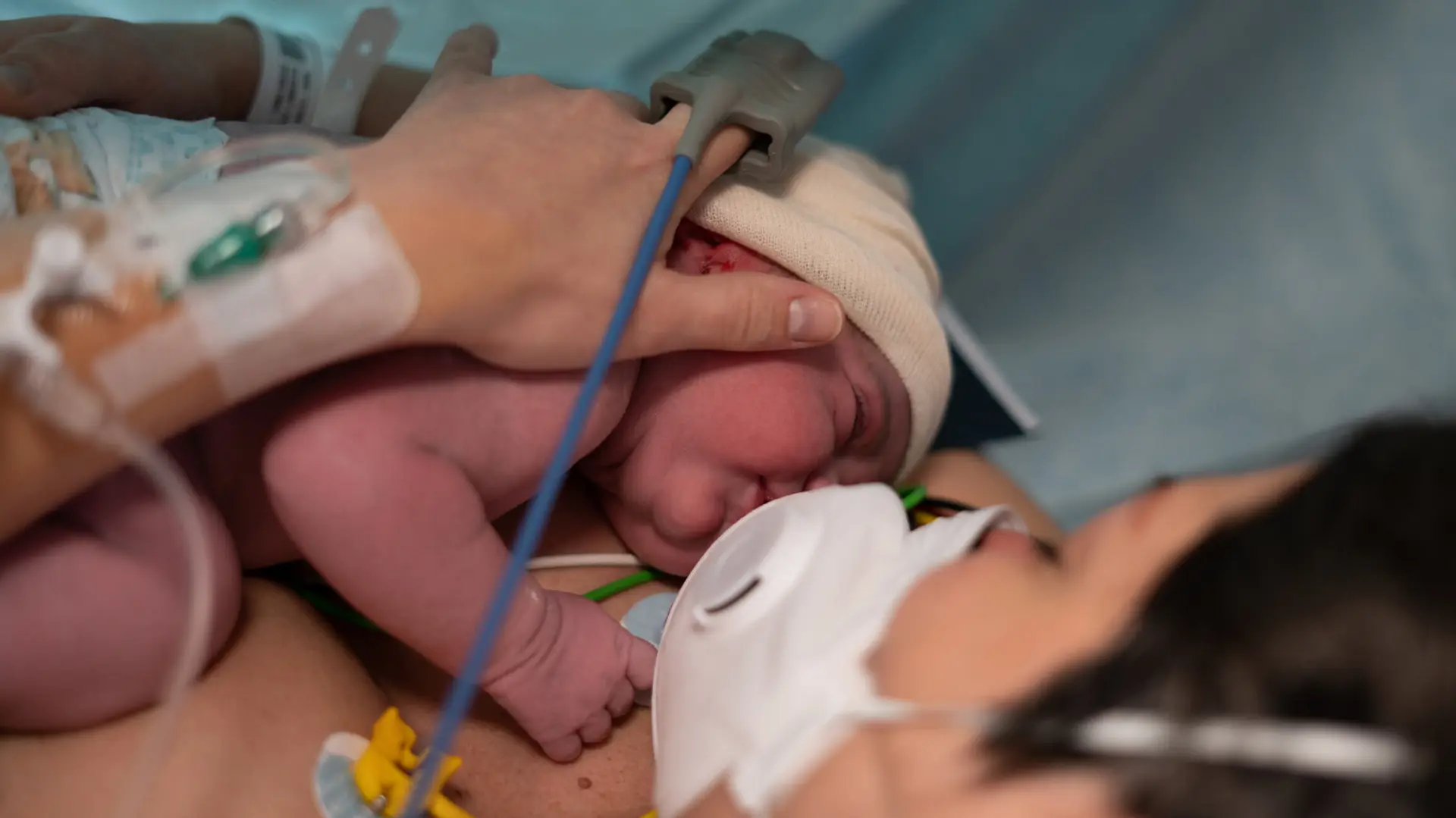 a mother lays in a hospital bed with a mask on and holds her newborn baby who may have contracted an infection against her chest