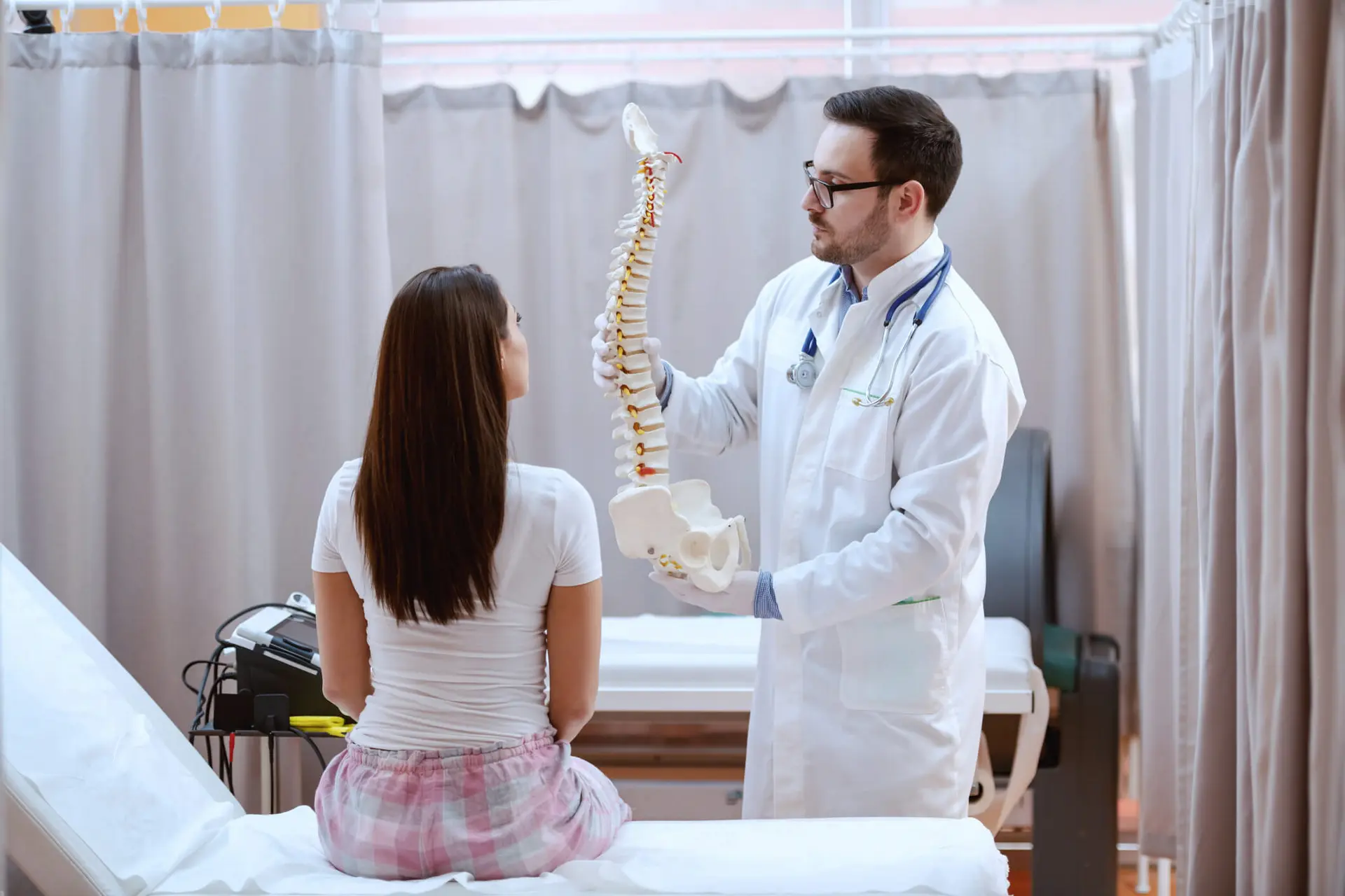 Young male Chiropractor in white uniform holding spine model and showing to his female patient.