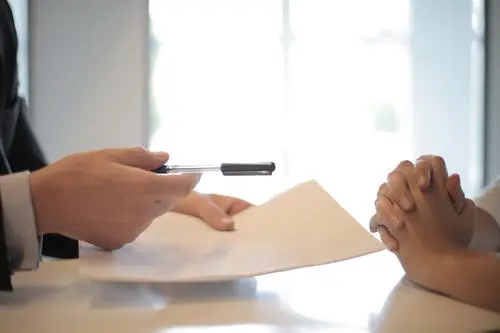 folded hands resting on table across from someone holding a pen and papers