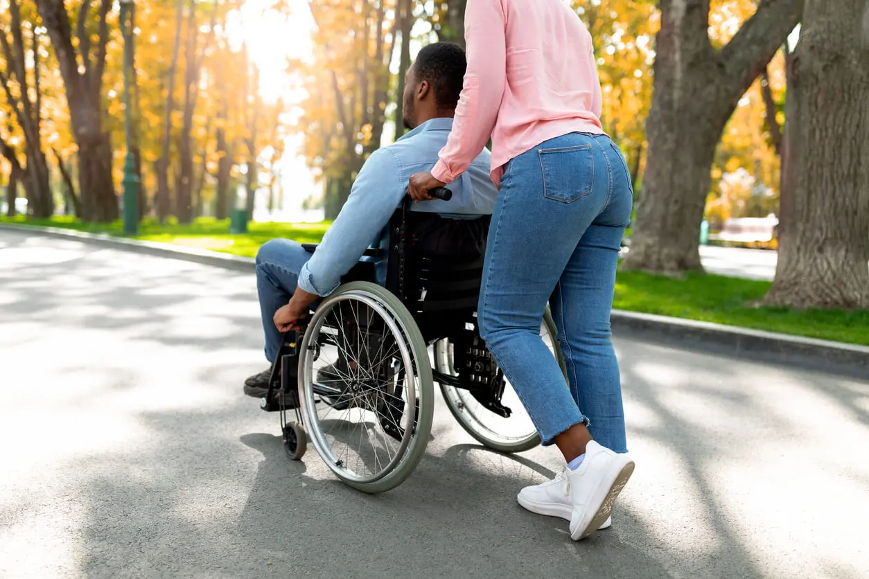 Young black woman pushing her husband who suffered a spinal cord injury in a wheelchair. Going for walk and enjoying the fall nature together outdoors.