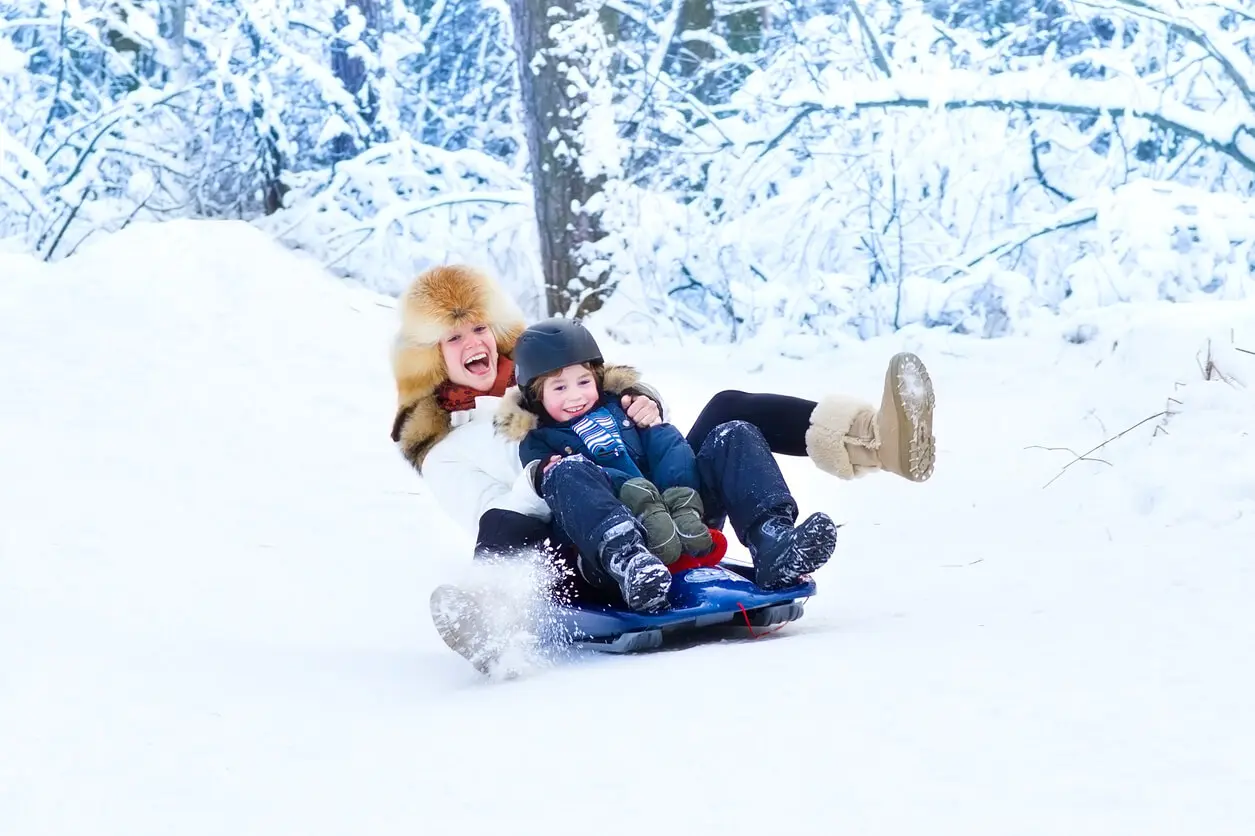 Young happy mother and her son wearing a helmet on a sleigh ride together in a snowy park