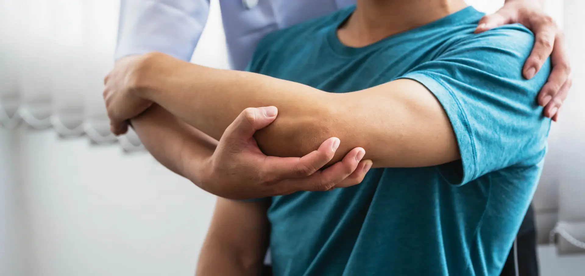 A male patient receives physical therapy treatment from a male physiotherapist at a clinic. The clinician stretches the patient's shoulder and arm. Photo Credit: iStock